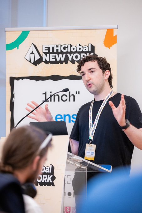 A man speaks at a podium during ETHGlobal New York, gesturing with his hands while a banner displays '1inch' in the background.