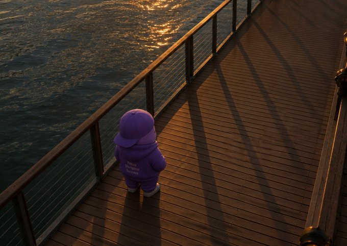 A 3D-rendered scene of a small character wearing a purple hoodie that reads "Young Dreams Success," standing on a wooden pier beside a railing overlooking sunlit water at golden hour.