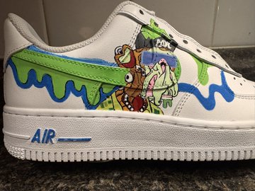 A close-up of a custom white Nike Air Force 1 sneaker featuring hand-painted artwork, including a green slime drip, characters from 'Aaahh!!! Real Monsters', and blue wavy lines.