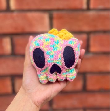 A hand holds a colorful, crocheted skull plushie with a yellow bow and large black eye sockets against a brick wall background.