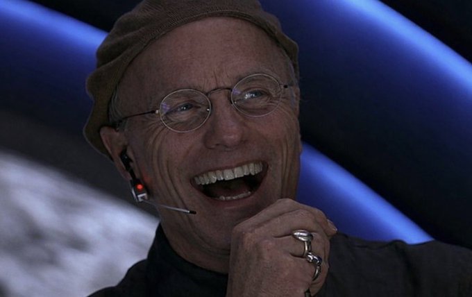 A man wearing a beanie, glasses, and a headset laughs joyfully against a blue and black background.