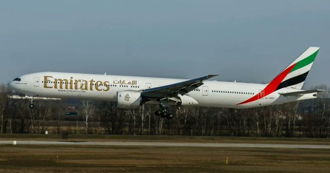 An Emirates Boeing 777 aircraft, featuring the airline's distinctive red, green, and white tail livery along with 'Emirates' branding in English and Arabic, is captured during landing with its landing gear deployed.