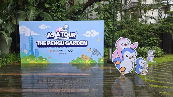 An outdoor event setup featuring a large illustrated sign reading 'ASIA TOUR TAIPEI EDITION - THE PENGU GARDEN' sponsored by Deepcoin and O.O Media, accompanied by cartoon penguin cutouts on a wet wooden deck surrounded by tropical plants.