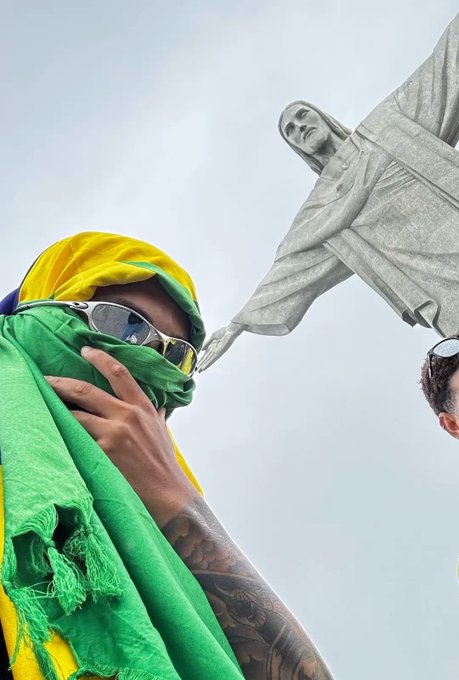 A low-angle photo showing a person wearing a green and yellow cloth/scarf over their face along with sunglasses, standing in front of a large stone statue of Christ with outstretched arms against a cloudy sky.