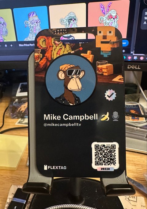 A FLEXTAG digital business card for 'Mike Campbell' (@mikecampbelltv), featuring a pixel-art monkey illustration and a QR code, displayed on a stand in front of a computer monitor.