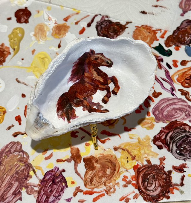 A small painted artwork of a rearing brown horse inside a white seashell, displayed on a paint-splattered surface.