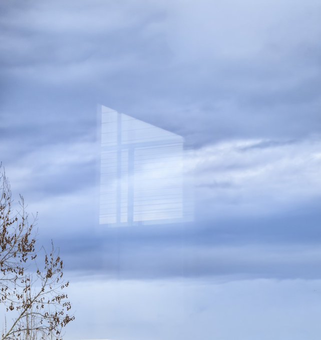 A reflection of a building's glass windows with horizontal blinds creates a ghostly overlay against a cloudy blue sky, with bare tree branches visible in the lower left corner.