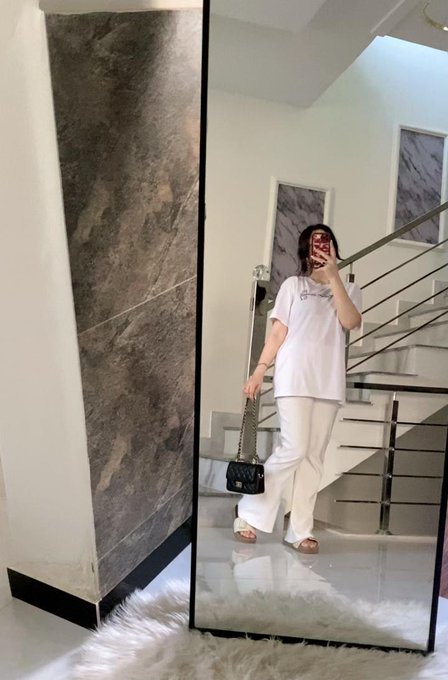 A woman takes a mirror selfie in an indoor setting, wearing a white t-shirt, white pants, and a shoulder bag, standing near a staircase.