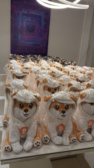 Rows of identical Shiba Inu dog plushies wearing hoodies with the Bitcoin ₿ symbol are individually wrapped in plastic.