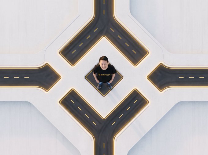 An aerial, top-down view of a person wearing a black 'BINANCE' t-shirt standing at the center of an illuminated cross-intersection where four roads meet, creating a symmetrical X-shaped pattern.