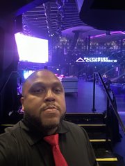 A man in a black shirt and red tie takes a selfie in what appears to be a gaming or streaming setup, with purple lighting and a "GAME FUEL" sign visible in the background.