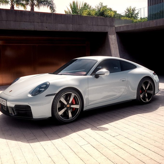 A silver Porsche 911 sports car parked in front of a modern building with palm trees visible in the background.