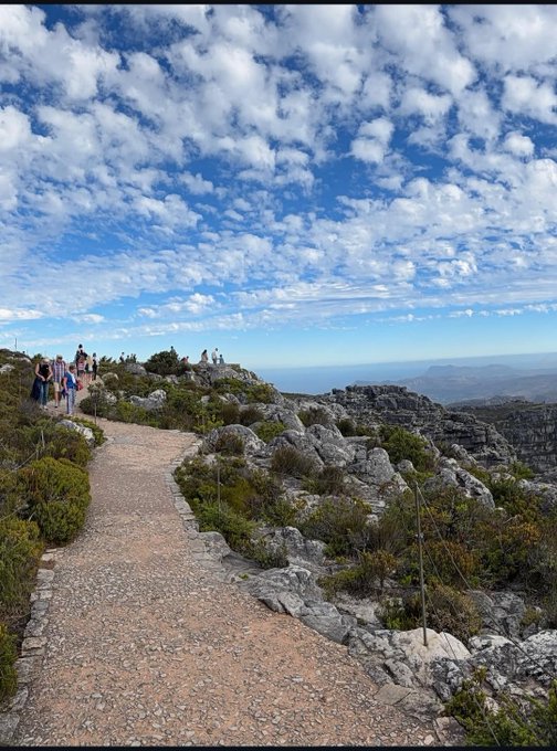 A scenic view of a rocky mountain path with people walking along it, set against a bright blue sky filled with scattered clouds and a distant landscape.