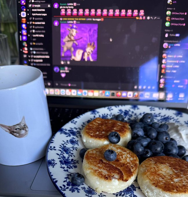 A cozy scene featuring a white mug with a cat face on it and a plate of small pancakes topped with blueberries in front of a laptop displaying a Discord chat with an AI-generated anime image.