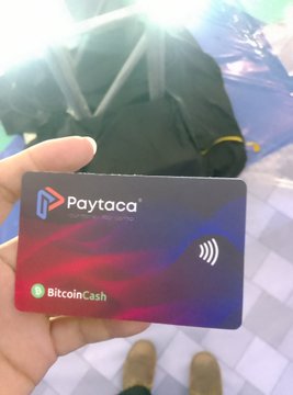A hand holds a black 'Paytaca' cryptocurrency card branded with 'BitcoinCash' and a contactless payment symbol, with a black bag visible in the background.