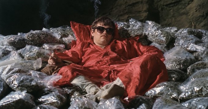 A scene from a film showing a man wearing sunglasses and a red outfit reclining on a large pile of plastic-wrapped packages.