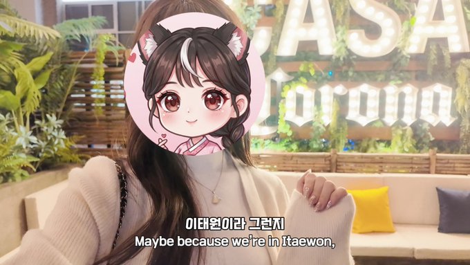 A selfie video of a woman with an anime cat-ear avatar overlay, sitting in a cafe with "ASIA" neon lights, subtitled "이태원이라 그런지 / Maybe because we're in Itaewon,"