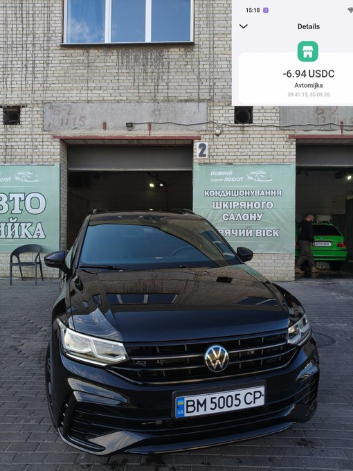 A black Volkswagen car parked outside an auto detailing shop with Ukrainian signage "КОНДІЦІОНУВАННЯ ШКІРЯНОГО САЛОНУ" (Leather Interior Conditioning), displaying a transaction notification of -6.94 USDC and license plate BM 5005 CP.