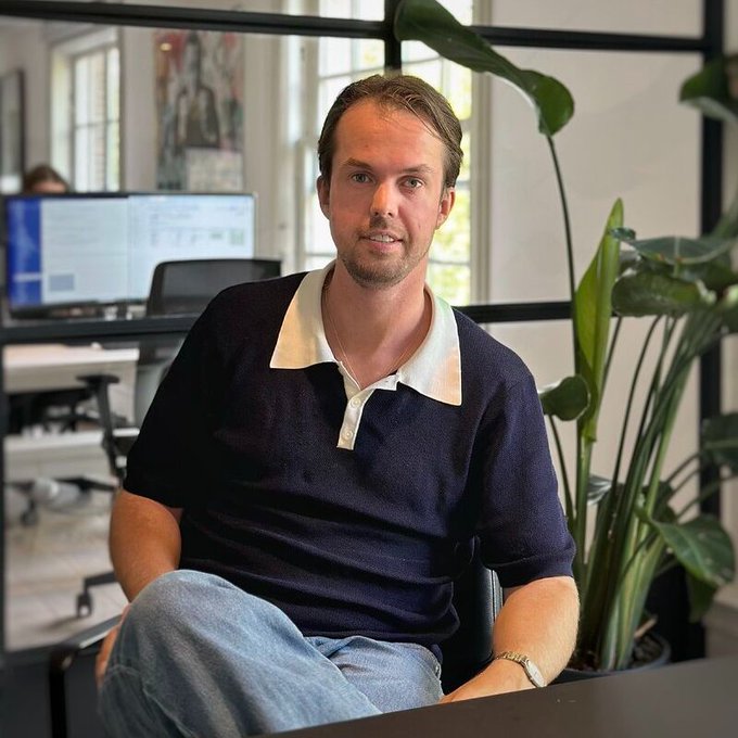 A man with short blonde hair and a beard wearing a navy blue polo shirt sits in an office setting with a plant and computer monitors in the background.