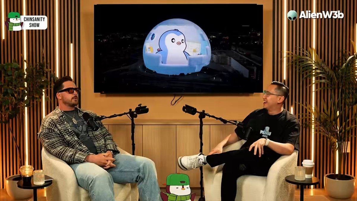 The image shows two men sitting in chairs, with a television screen behind them displaying an animated penguin character. The man on the left side of the image appears to be wearing glasses and has his arms crossed over his chest. The other man is seated comfortably with his legs crossed. Both men seem to be engaged in conversation or watching something on the TV screen together.