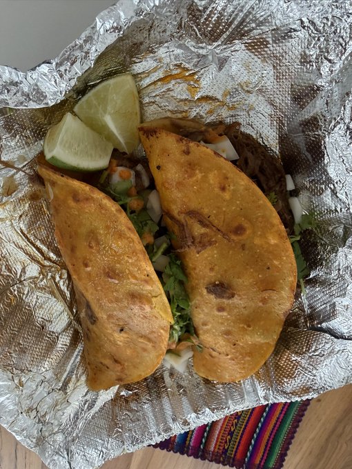 Two golden-brown crispy tacos filled with lettuce, onions, and meat are served on foil alongside lime wedges, capturing a casual, flavorful meal that was enjoyed today. The vibrant Mexican-style dish sits atop a colorful striped cloth, adding to the homemade, authentic presentation.