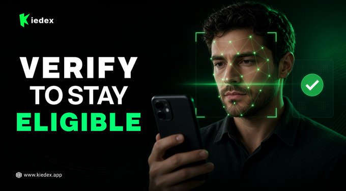 The image is a promotional graphic featuring a man looking at his phone with a facial recognition scan overlaid on his face, accompanied by the bold text "VERIFY TO STAY ELIGIBLE" and the KieDex logo with the URL www.kiedex.app.

The graphic visually reinforces the post's announcement about KieDex's mandatory KYC verification, using facial recognition imagery to symbolize the new identity verification requirement aimed at eliminating fake and inactive accounts from the ecosystem.