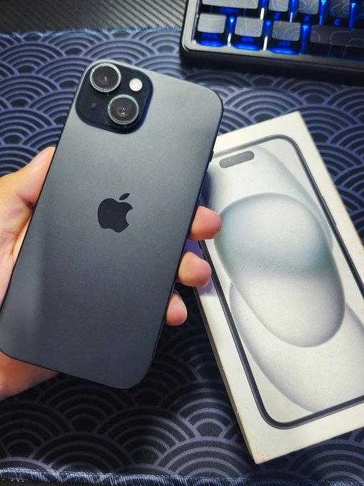 A hand holding a black iPhone (appears to be an iPhone 13 mini or similar) showing the back with the Apple logo and dual camera setup, with its white product box beside it on a blue wave-patterned mat, with a blue-backlit mechanical keyboard visible in the background.