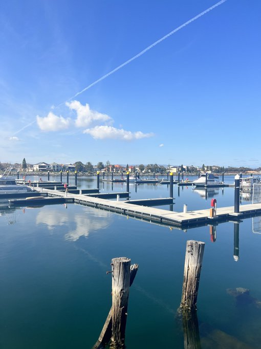 A serene marina scene with wooden docks, boats, and glassy water under a blue sky with scattered clouds.