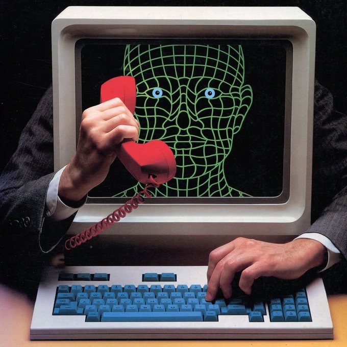 A person in a suit holds a red telephone receiver to a vintage computer monitor displaying a wireframe face, typing on the keyboard—evoking themes of communication, digital interaction, and technology that complement the post's announcement of Variational's upgraded trading platform UI.
