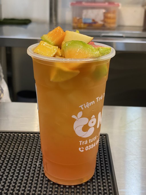 A plastic cup of fruit tea with diced mango and what appears to be starfruit is topped on a bar counter, branded with a 'T' logo and a phone number.