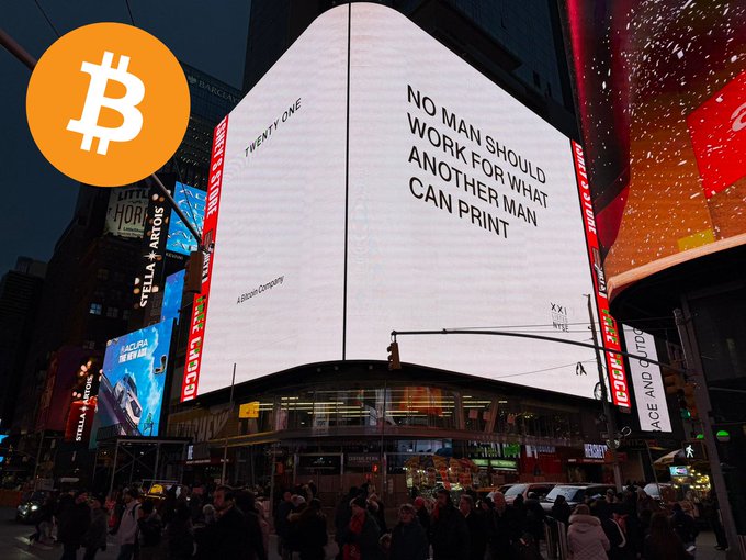 Nighttime photograph of a brightly lit billboard in Times Square displaying the Bitcoin logo and the text 'NO MAN SHOULD WORK FOR WHAT ANOTHER MAN CAN PRINT,' alongside references to Twenty One and a Bitcoin company.