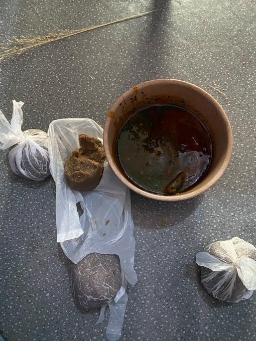 A top-down view of a street food meal on asphalt, featuring a bowl of red soup or curry, some bread or fried food on plastic wrap, and two small plastic bags containing what appears to be additional food items.