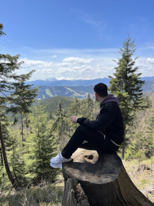 A person wearing a black jacket and white sneakers sits on a tree stump at the top of a mountain, looking out over a forested landscape with snow-capped peaks in the distance.