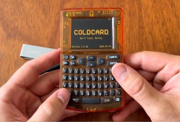 A pair of hands holds a transparent Coldcard hardware wallet displaying the "COLDCARD" logo, version 1.5.12, dated 2025-01-15 on a wooden surface—visually reinforcing the urgent warning about entropy-related vulnerabilities in Coldcard Mk3 wallets.