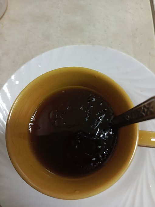 A top-down view of a yellow ceramic cup filled with black coffee or tea, containing a metal spoon and sitting on a white saucer.