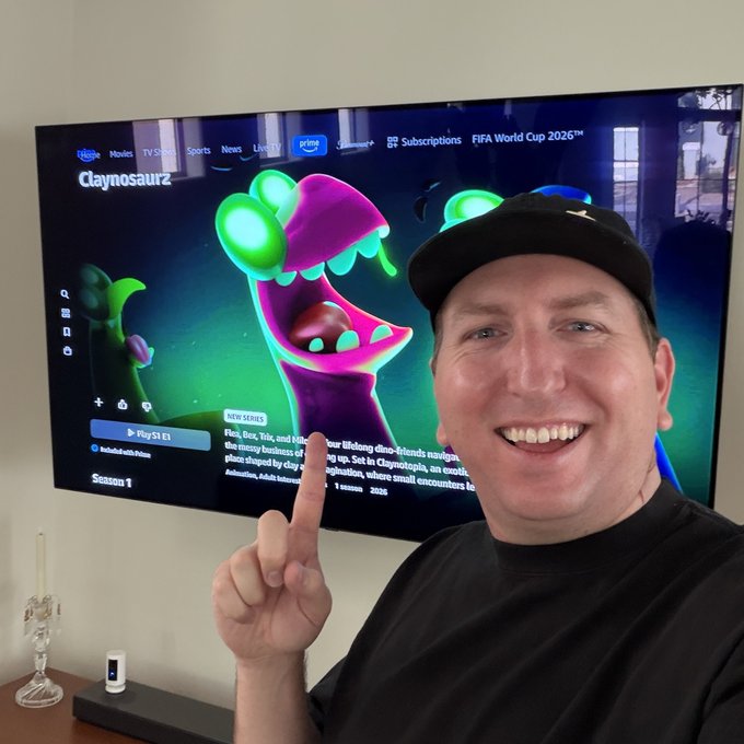 A smiling man in a black cap pointing at a TV screen showing the 'Claynosaurz' animated series on Prime Video.