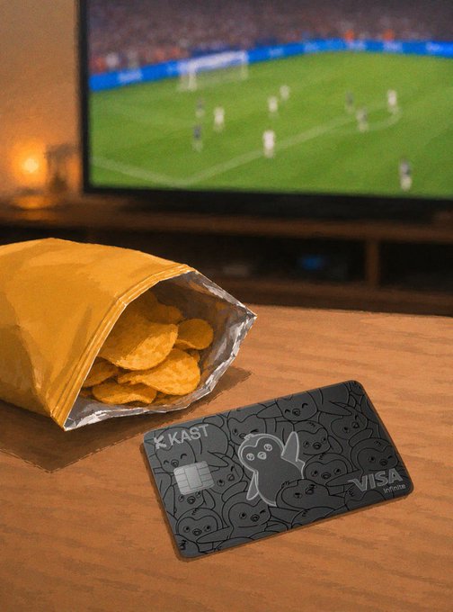 A KAST Visa debit card next to an open bag of chips on a wooden coffee table, with a soccer/football match playing on a TV in the background.