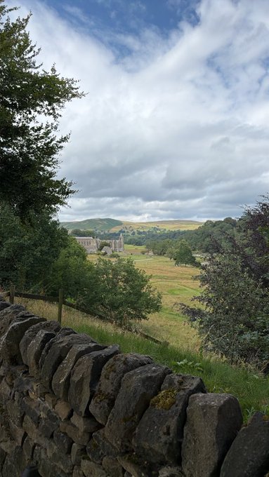 A scenic view of the Yorkshire Dales at dusk, with a stone wall in the foreground framing rolling green hills and what appears to be a historic abbey ruins in the distance under a dramatic cloudy sky. The landscape perfectly complements the "Good Evening from the Yorkshire Dales" greeting, capturing the area's iconic blend of pastoral countryside and historical landmarks.