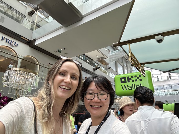 Two smiling women take a selfie at an OKX Wallet booth located inside a brightly lit shopping mall, with a FRED store visible in the background.