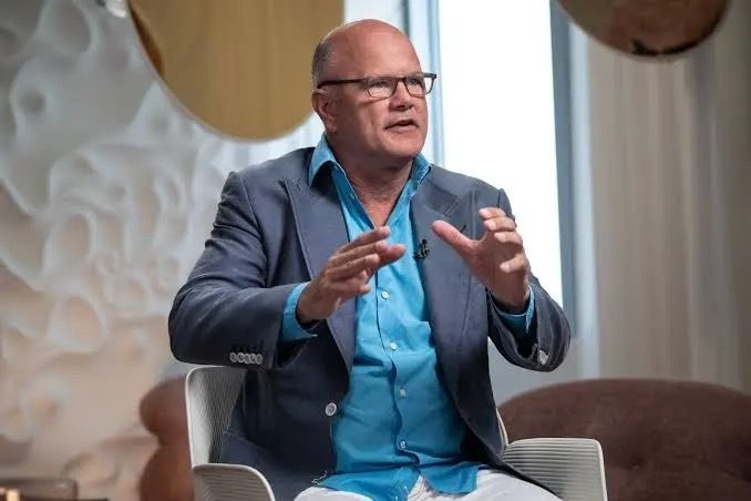 Mike Novogratz, wearing glasses and a blue shirt under a gray blazer, is shown mid-gesture during an interview. The image relates to the post by visually representing Novogratz, the prominent crypto advocate quoted discussing the CLARITY Act's potential revival.