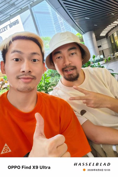 A selfie of two men, one in an orange shirt giving a thumbs up and another in a white t-shirt making a peace sign while wearing a bucket hat, watermarked with 'OPPO Find X9 Ultra' and 'HASSELBLAD'.