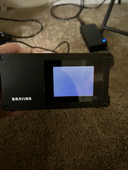 A hand holds a black 'BRAIINS' branded Bitcoin mining device featuring a small display screen, connected via cables on a carpeted surface.