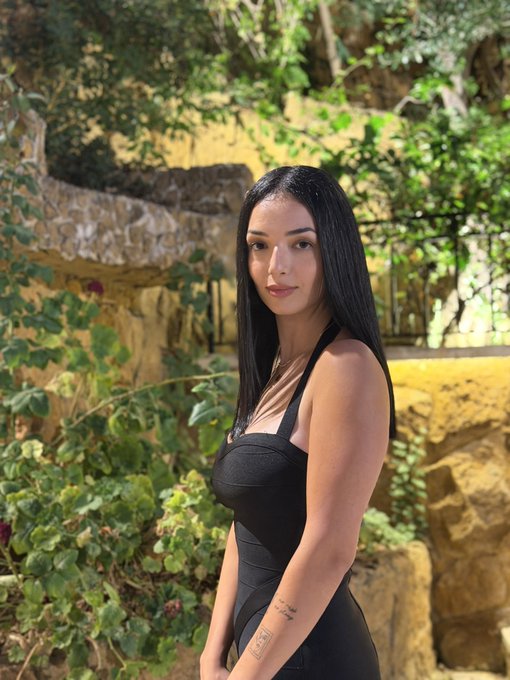 A young woman with long dark hair poses in a black halter top and bottoms against a backdrop of stone walls and lush greenery in a garden setting.