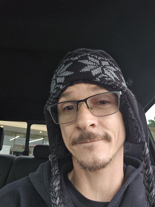 A selfie of a man wearing glasses, a mustache, and a patterned knit hat with ear flaps inside a car.