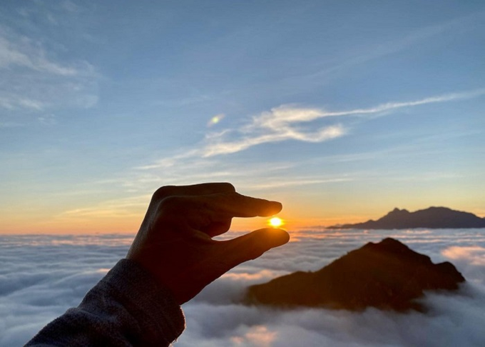 A silhouette of a hand pinching the sun between thumb and forefinger above a sea of clouds at sunset, with a mountain peak visible in the distance.