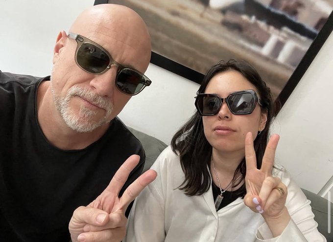 The image features a man wearing sunglasses and a woman posing with her fingers crossed, both of them smiling for the camera. They are sitting next to each other, possibly at an event or gathering. The background includes a painting hanging on the wall behind them. The caption "A ghost report on Zero 10 coming soon" suggests that there might be some sort of event or gathering taking place in this location, and it is likely related to the post about the Ghost Report.