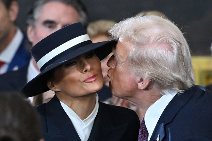 Donald Trump kisses Melania Trump on the cheek; she wears a black wide-brimmed hat with a white band and a black blazer, while he wears a navy suit.