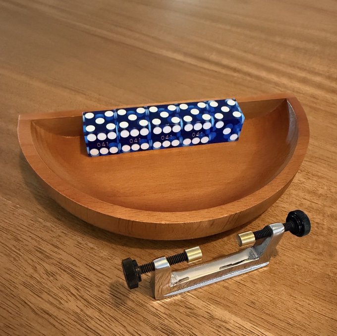 A wooden dice tray containing a set of four blue dice with white pips, accompanied by two metal dice clamps with brass and black knob components, all resting on a light wood-grain surface.