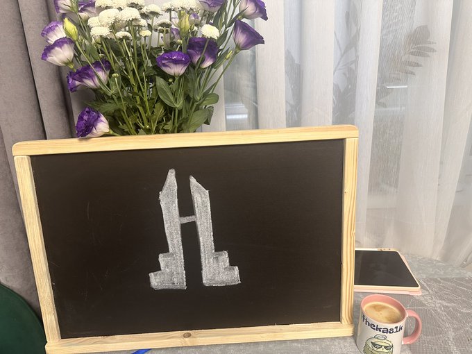 A small wooden-framed chalkboard displays a silver glittery letter 'H', sitting next to a vase of purple and white flowers, a smartphone, and a pink mug with 'theMask' written on it.