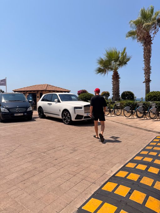A man in a red cap and black outfit walks across a brick driveway toward a white Rolls-Royce Cullinan parked near palm trees and bicycles, setting the scene for a luxurious "Gm friends" greeting. The coastal, sunlit location with its upscale vehicles complements the casual, friendly vibe of the post.